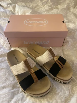 Graceland sandaler med flätad sula prop - Snygga sandaler från Graceland i storlek 39/UK6. De har breda remmar i beige tyg och svart mockaimitation, samt bruna detaljer med guldfärgade ringar. Sulan är flätad i espadrillestil och innersulan är mjuk och stripad i beige. Perfekta för en trendig och avslappnad look.