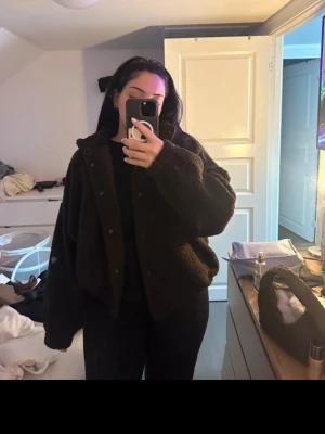 Mörkbrun teddyjacka med hög krage - Mysig mörkbrun teddyjacka med hög krage och tryckknappar framtill. Jackan har två sidofickor och är tillverkad i fluffigt teddy-material som ger en avslappnad och trendig look. Perfekt för att hålla sig varm under kyliga dagar.