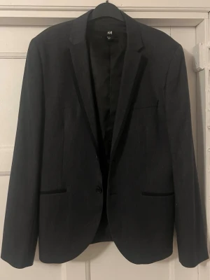 Svart kavaj från H&M - Stilren svart kavaj från H&M med klassisk passform och enkla slag. Kavajen har två fickor framtill och en knappstängning. Perfekt för en dressad look och funkar lika bra till skjorta som t-shirt.