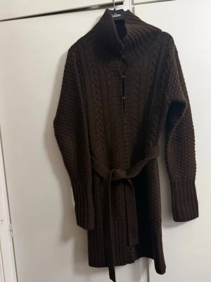 Mörkbrun stickad kofta med skärp - Mysig mörkbrun stickad kofta med grovt kabelstickat mönster, hög krage och knappar framtill. Koftan har ett matchande midjeskärp och långa ärmar med ribbade muddar. Perfekt för lager på lager och höstmys.