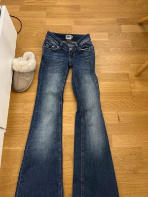 Blå bootcut jeans från 157 - Säljer ett par blå bootcut jeans från 157 med klassisk femficksdesign och snygga slitningar framtill. Jeansen har låg midja och utsvängda ben, perfekt för en trendig och avslappnad look. Materialet är denim i bomull.