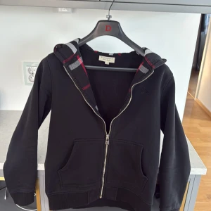 Svart hoodie från Burberry med dragkedja - Svart hoodie från Burberry med hel dragkedja och klassiskt rutigt foder i huvan. Två fickor framtill och broderad logga på bröstet. Perfekt för en stilren och avslappnad look.