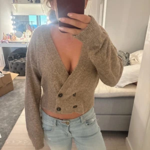 Beige croppad kofta med knappar - Säljer en beige croppad kofta med djup v-ringning och dubbla rader svarta knappar framtill. Koftan är stickad och har långa ärmar med breda muddar. Perfekt för lager-på-lager och chill vardagsstil.