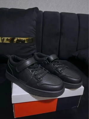 Svarta skor - Snygga helsvarta sneakers med både snörning och kardborreband. Skorna har en klassisk låg profil, perforerad tå och är tillverkade i konstläder för en clean look. Perfekta för dig som gillar stilrena och praktiska skor.