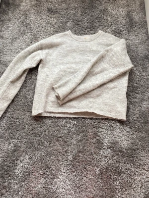 Ljusgrå stickad tröja Gina Tricot - Mysig ljusgrå stickad tröja från Gina Tricot med rund halsringning och långa ärmar. Tröjan har en enkel, klassisk design och är tillverkad i mjukt material som passar perfekt till kalla dagar.