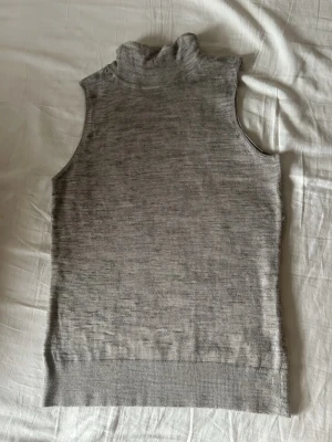 Grå ärmlös polotopp i ull - Snygg grå ärmlös polotopp i mjuk ull. Toppen har hög krage och ribbad kant nedtill, perfekt för lager på lager och en clean look. Passar till både jeans och kjol för en stilren outfit.