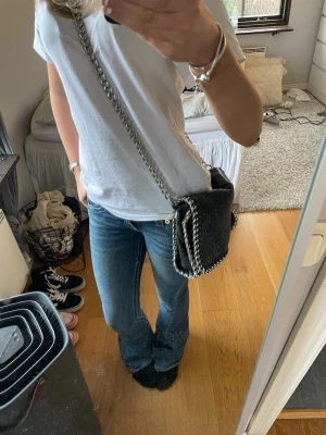 Svart tiamo väska axelväska - Säljer en svart axelväska från Tiamo med kedjerem och silverfärgade detaljer. Väskan har en mjuk, vegansk läderlook och kantas av en snygg kedja runt hela väskan. Perfekt storlek  till din outfit.