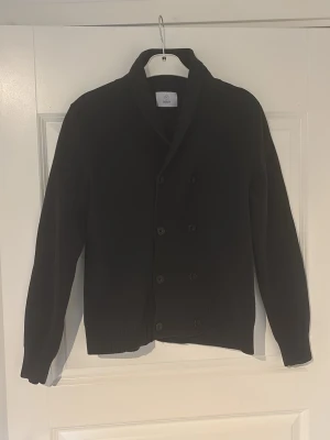 Svart dubbelknäppt kofta från BLK DNM - Stilren svart kofta från BLK DNM med dubbelknäppning framtill och bred krage. Koftan har långa ärmar och ribbade muddar vid ärmslut och nederkant. Perfekt för lager på lager och en clean look. Ursprungligen storlek L, men sitter som S/XS, vet ej om det är modellen eller att den har krympt.