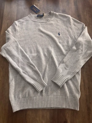 Ljusgrå stickad tröja Polo Ralph Lauren - Snygg ljusgrå stickad tröja från Polo Ralph Lauren med rund halsringning och diskret blå logga broderad på bröstet. Tröjan har långa ärmar och ribbade muddar vid ärmslut och nederkant. Perfekt till jeans eller chinos.