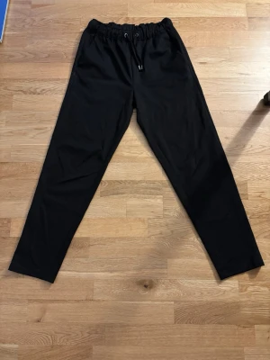 Svarta kostymbyxor - Säljer ett par svarta mjukisbyxor med dragsko i midjan och raka ben. Byxorna har en avslappnad passform och är perfekta för chill dagar. Materialet känns mjukt och bekvämt, troligen bomull eller bomullsblandning. Inga synliga interface eller loggor.