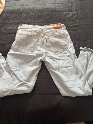 Levi's 501 ljusblå raka jeans - Klassiska Levi's 501 jeans i ljusblå denim med raka ben och hög midja. Jeansen har två bakfickor med den ikoniska sömmen och röd Levi's-tag. Tillverkade i bomull för en skön och tidlös look.