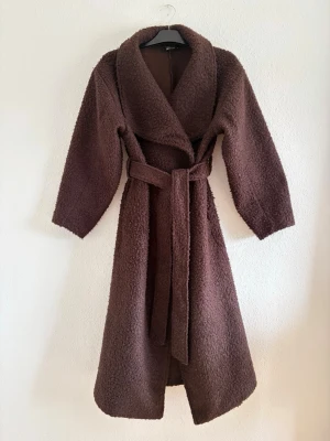 Soft boucle long coat - Mysig brun teddy kappa med bred sjalkrage och knytskärp i midjan. Lång modell med avslappnad passform och långa ärmar. Perfekt för kalla dagar och ger en trendig look. Tillverkad i mjukt, fluffigt material som känns riktigt skönt.