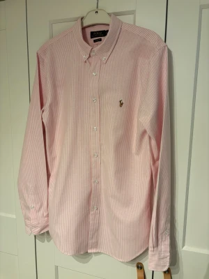 Rosa randig skjorta från Polo Ralph Lauren - Klassisk rosa och vit randig skjorta från Polo Ralph Lauren med button-down krage och broderad logga på bröstet. Skjortan har långa ärmar och knäppning framtill. Perfekt för en clean och stilren look. Knappt använd!