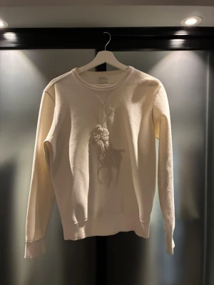 Vit sweatshirt från Polo Ralph Lauren - Snygg vit sweatshirt från Polo Ralph Lauren med broderad logotyp på bröstet. Tröjan har rund halsringning, långa ärmar och ribbade muddar. Perfekt för dig som gillar klassisk och stilren design.