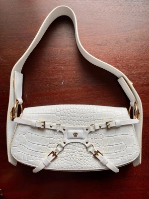 Manc Lilith croc med gulddetaljer - Influencer / designerväska från Manc i 100% äkta läder. Originalpris är ca 5300kr utan tullavgifter. I modellen Lilith. Axelbandet går att förkorta. Måtten är: 5 cm (djup) 🌸 26 cm (längd) och 10 cm (höjd om man exkluderar förslutningen). Annars är den totala höjden 14 cm🌸 Det finns slitningar på vissa av gulddetaljerna och vissa missfärgningar (ej något som är särskilt synligt vid användning). 