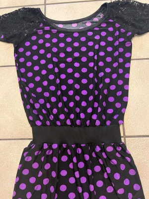 Svart klänning med lila prickar storlek S/M - Svart kortklänning med lila prickigt mönster och bred resår i midjan. Klänningen har korta spetsärmar och rund halsringning. Perfekt för dig som gillar färg och mönster!