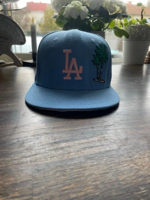 Blå LA Dodgers keps med palmer - Snygg ljusblå LA Dodgers keps från New Era med broderad LA-logga i rosa och palmmotiv på framsidan. Klassisk flat brim och MLB-logga bak. Tillverkad i bomull för en bekväm känsla och stilren look.