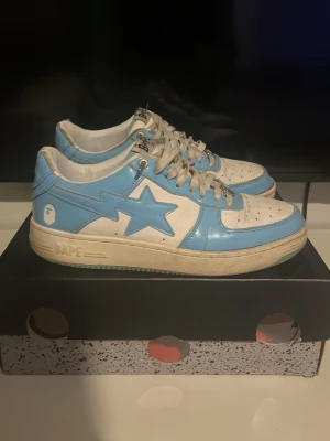 BAPE STA blå och vita sneakers - Säljer ett par BAPE STA sneakers i blått och vitt läder med den ikoniska stjärnan på sidan. Skorna har vita snören, perforerad tåbox och BAPE-logga på hälen. Klassisk low-top siluett som sticker ut med sin färgkombination och streetwear-vibe.