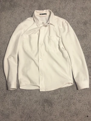 Vit skjorta från Zara med fickor - Stilren vit skjorta från Zara med två stora bröstfickor och klassisk krage. Skjortan har långa ärmar och knäppning framtill. Perfekt för en clean och modern look.