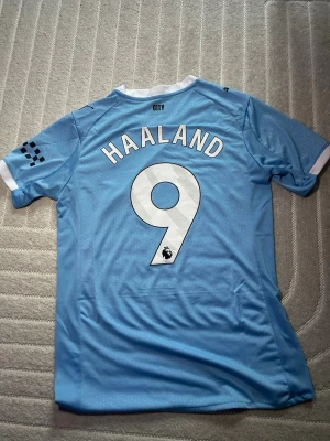 Manchester City Haaland matchtröja - Snygg ljusblå Manchester City matchtröja från Puma med HAALAND och nummer 9 på ryggen. Tröjan har klubbmärke och sponsortryck på bröstet samt vita och mörkblå detaljer. Tillverkad i lätt och ventilerande funktionsmaterial, perfekt för fotbollsträning eller match.