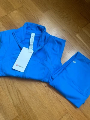 Blå träningsset från Lululemon - Snyggt blått träningsset från Lululemon med dragkedja på tröjan och matchande tights. Setet är gjort i ett stretchigt och mjukt funktionsmaterial som passar perfekt till gymmet eller löprundan. Diskret logga på byxan och tröjan har hög krage.