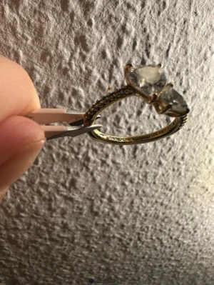 Guldfärgad ring med hjärtformade stenar - Pris kan vi diskutera privat. Snygg guldfärgad ring från Pandora med två stora hjärtformade klara stenar framtill och små gnistrande stenar längs bandet. Ringen har en elegant och romantisk design som verkligen sticker ut. Perfekt för dig som gillar detaljer och vill ha något extra.