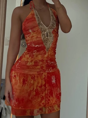 Orange halterneck klänning med pärlor - Säljer en kort halterneck klänning i orange och röda toner med blommigt mönster. Klänningen har pärldetaljer och djup v-ringning framtill samt omlottkänsla i kjolen. Materialet känns lätt och flowy, perfekt för varma dagar.