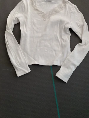 Vit ribbad långärmad topp med spets - Snygg vit ribbad topp med lång ärm och v-ringning dekorerad med spetsdetalj. Perfekt att styla till jeans eller kjol för en clean look. Mjuk och stretchig känsla.