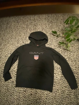 Svart GANT hoodie - Snygg svart hoodie från GANT med stor huva och klassisk logga på bröstet. Framtill finns GANT-text och ett broderat sköldmärke i rött, vitt och blått. Hoodien har långa ärmar och ribbade muddar. Perfekt för en avslappnad och stilren look.