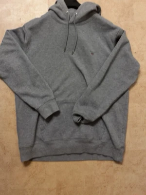 Gant hoodie - Säljer en klassisk grå hoodie från Gant med justerbar huva och känguruficka framtill. Diskret Gant-logga på bröstet och snygga metalldetaljer på snörena. Perfekt för dig som gillar stilren och bekväm streetwear.