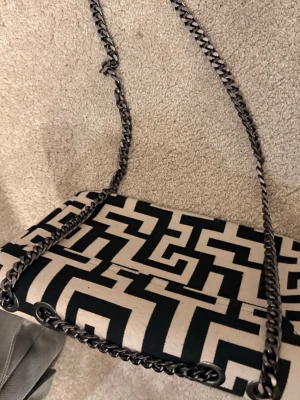 Svartvit axelväska från River Island - Säljer en snygg axelväska från River Island med svartvitt grafiskt mönster och kedjeremmar i metall. Väskan har rektangulär form och innerfack med dragkedja. Perfekt för dig som gillar statement-accessoarer och vill sticka ut.