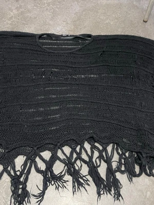 Svart virkad strandponcho med fransar - Snygg svart strandponcho i virkad design med stora maskor och dekorativa fransar längs nederkanten. Perfekt att slänga över bikinin på stranden för en avslappnad och trendig look. Materialet känns lätt och luftigt.