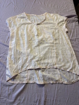 Vit broderad blus med spetsdetaljer - Söt vit blus med korta ärmar och broderade blommor. Blusen har rund halsringning, spetsdetaljer längs kanterna och en något längre bakdel. Tillverkad i ett tunt, luftigt tyg som känns perfekt för varma dagar. Finns ingen storlek i men skulle tro att det är L/XL