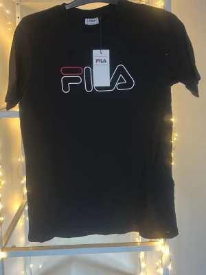 Svart FILA t-shirt med logga - Svart t-shirt från FILA med stor, ikonisk logga i vitt och rött på bröstet. Klassisk rund halsringning och korta ärmar. Tillverkad i mjuk bomull som känns skön mot huden. Perfekt för dig som gillar sportiga och stilrena plagg.