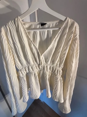 Vit plisserad blus med v-ringning - Säljer en vit plisserad blus med v-ringning och resår i midjan som ger en snygg siluett. Blusen har långa ärmar och ett vågigt strukturmönster i tyget. Perfekt för dig som gillar trendiga detaljer och en luftig känsla.