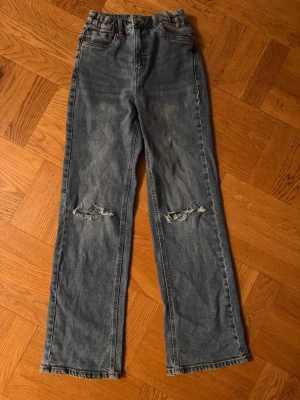 Blå raka jeans med slitningar - Säljer ett par blå raka jeans med slitna detaljer över knäna. Jeansen har hög midja, klassisk femficksmodell och är tillverkade i denim. Perfekta för en avslappnad och trendig look.