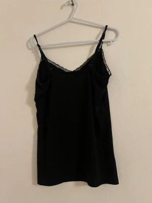 Svart linne med spetskant - Snyggt svart linne med tunna axelband och spetsdetalj längs urringningen. Perfekt att styla lager på lager eller bära som det är. Mjuk och stretchig känsla, troligtvis i bomull eller viskosblandning.