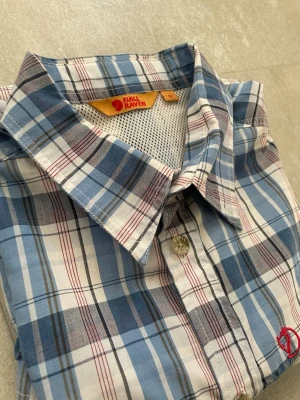 Blårutig skjorta från Fjällräven - Kortärmad skjorta från Fjällräven i blått, vitt och rött rutigt mönster. Skjortan har klassisk krage, knappar framtill och en liten bröstficka med röd Fjällräven-logga. Tillverkad i lätt och luftig bomull, perfekt för varma dagar.