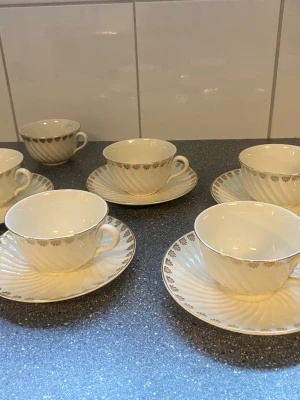 6 vita kaffekoppar med fat och guldkant - Uppsala-Ekeby Gefle / Säljer ett set med 5 äggskalsvita kaffekoppar och tillhörande fat i porslin. Kopparna och faten har ett elegant snirklat mönster och dekoreras av en tunn guldkant samt små guldfärgade detaljer längs kanten. Perfekt för dig som gillar klassisk och stilren design.