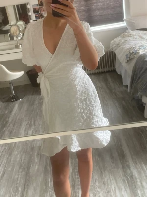 Vit omlottklänning med volang - Söt vit omlottklänning med volangkant och knyt i midjan. Klänningen har korta puffärmar och ett diskret blommigt mönster i tyget. Perfekt för studenten!