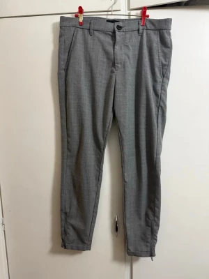 Grå byxor med smal passform från GABBA - Snygga grå byxor , modern stadslook, med en avsmalnande passform och de ikoniska Gabba Pisa-dragkedjorna vid bensluten. Med den stiliga uppvikta fållen, ett högstretchigt bomullsblandat tyg och diskreta kritstreck får du en perfekt metropol-stil för alla tillfällen. De perfekta byxorna – oavsett om du klär upp dig eller satsar på en mer avslappnad look