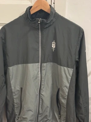  träningsjacka Försvarsmakten (M) - Snygg tvåfärgad grå vindjacka i fint skick (Storlek M) från Trimtex med dragkedja framtill, hög krage och två fickor med dragkedja. Jackan har logga på bröstet och texten 'FÖRSVARSMAKTEN' på ryggen. Tillverkad i lätt och vindtätt material, perfekt för blåsiga dagar. Ena spännbanden har öppnat upp sig, går enkelt att fixa och det är varken något som syns eller påverkar jackan. 