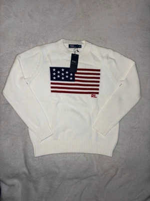 Vit stickad tröja från Polo Ralph Lauren - Vit stickad tröja från Polo Ralph Lauren med amerikanska flaggan broderad på bröstet och RL-logga i rött. Tröjan har rund hals, långa ärmar och ribbstickade muddar. Perfekt för dig som gillar klassisk och stilren design. Ärmlängd:60 längd: 59 bröst: 48 axelbredd: 36