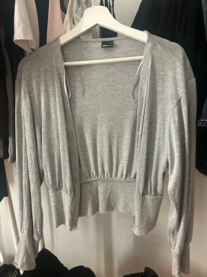 Grå cropped kofta från Gina Tricot - Säljer en ljusgrå, tunn och mjuk cropped kofta från Gina Tricot. Koftan har långa ärmar, öppen front och resår i midjan för en snygg passform. Perfekt att slänga över ett linne eller t-shirt för en chill look.