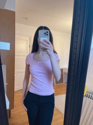 Ljusrosa topp med rosett och spets - Söt ljusrosa topp med fyrkantig ringning, dekorerad med en liten rosett och spetskant. Kortärmad och figurnära passform, perfekt för dig som gillar en feminin och enkel stil. Materialet känns mjukt och stretchigt, troligen bomull eller syntet.