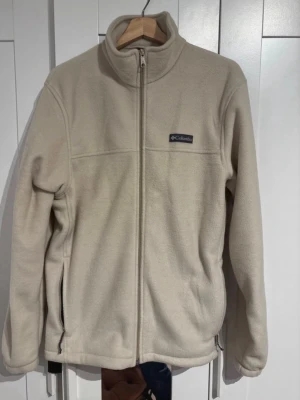 Beige fleecejacka från Columbia - Mysig beige fleecejacka från Columbia med hel dragkedja och hög krage. Jackan har två sidofickor med dragkedja och en diskret logga på bröstet. Perfekt för lager-på-lager och sköna höstdagar.