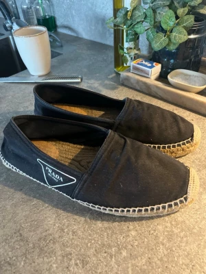 Svarta Prada loafers i tyg - Säljer ett par svarta loafers från Prada med klassisk espadrille-look. Skorna är i tyg med naturfärgad flätad sula och har en tydlig Prada Milano-logga på sidan. Perfekta för en avslappnad men ändå stilren look.