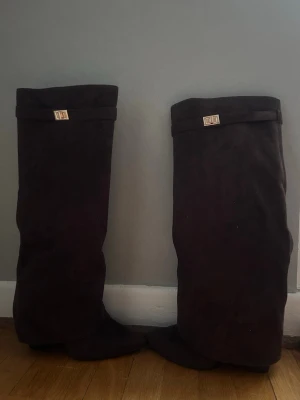 Mörkbruna mockastövlar med gulddetalj - Snygga mörkbruna stövlar i mocka med spetsig tå och dekorativ gulddetalj upptill. Stövlarna har en hög och slouchy design som ger en cool och trendig look. Perfekta att styla med både jeans och kjol. Säljer pga att klacken var lite för hög för mig! Har endast använt bootsen en gång men på den vänstra skon är det lite slitage vid tån. Nypris 1000