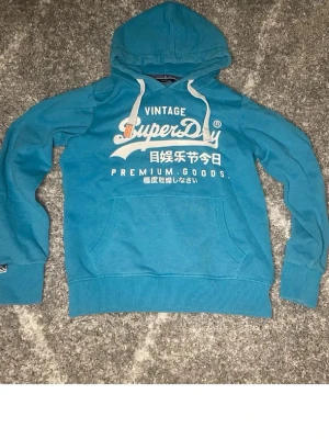 Blå Superdry hoodie med tryck - Snygg blå hoodie från Superdry med vit logga och text framtill. Klassisk känguruficka, ribbade muddar och vit dragsko i huvan. Perfekt för en avslappnad och trendig stil. Mjuk bomullsblandning som känns skön mot huden.