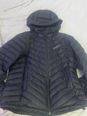 Svart dunjacka med huva PeakPerformance - Snygg svart dunjacka från PeakPerformance med huva och dragkedja framtill. Jackan har ett quiltat mönster, två sidofickor med dragkedja och logga på bröstet. Perfekt för kalla vinterdagar tack vare det lätta och värmande dunmaterialet.